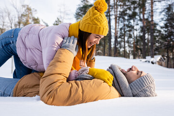 Side view of cheerful man lying on snow near girlfriend in winter outfit