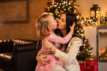 Obraz na płótnie Canvas Pretty young mother woman with little daughter girl in fashionable clothes kiss on the background of christmas decorations and lights. Family winter holidays