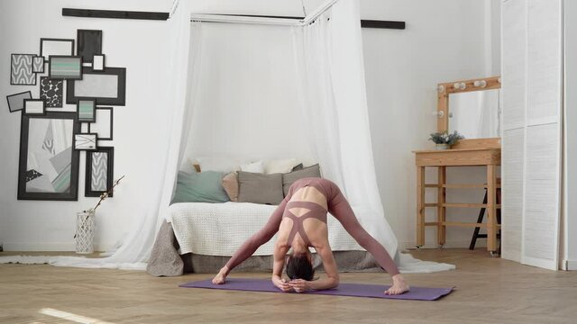 Slender, flexible, caucasian middle-aged woman with black hair in beige tracksuit performs standing asanas Prasarita Padottanasana, Wide-Legged Forward Fold on purple yoga mat in bright room with bed