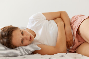 Portrait of sick woman wearing white casual t shirt lying in bed, touching her belly, suffering from terrible abdominal pain, posing indoor, health problems.