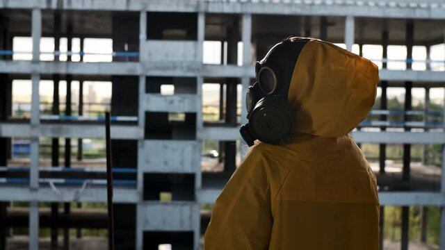 Human in gas mask and yellow protective suit sits in abandoned building, looks into distance and turns his face to us. Concept of toxic loneliness, post-apocalyptic world and disappointment.