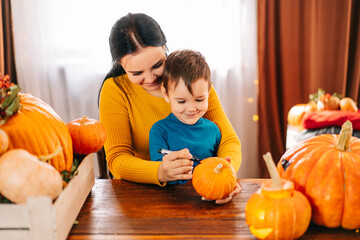Fototapeta premium A happy family is getting ready for Halloween. a beautiful young mother and her son draw a pumpkin's face at home with a black marker.