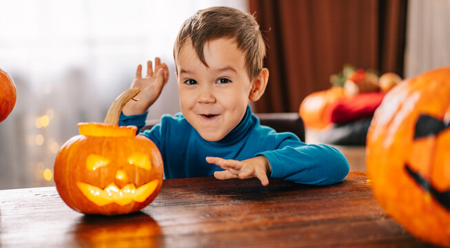 A Beautiful Little Boy Carves A Pumpkin And Smiles, On A Background Decorated For Halloween. The Boy Is Sitting At The Table And Making Faces