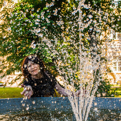 Senior happy woman by the fountain catches water droplets on a sparkling sunny summer day.