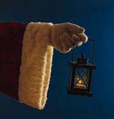 Hand of Santa Claus in a white glove holding a lantern with a burning fire, in the dark. Blue night background, fire, sacrament, winter. Santa Claus illuminates the road in the dark.