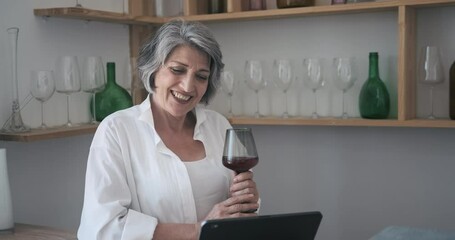 Senior old attractive woman using tablet for video call, drinking wine and participating in a chat with children, participating in family distant party celebration, active leisure feeling alone - Powered by Adobe