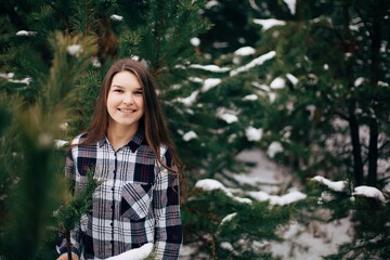Portrait of a beautiful girl with long hair in a snowy forest. Winter in the woods. Girl walks in the winter forest. Checkered shirt. Cold winter day. Beautiful pines in the snow