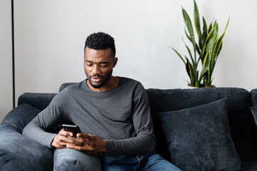 African-American guy sitting on the couch and using smartphone, chatting in social networks, texting