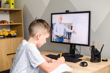 Schooler boy is studying in front of the computer, taking a part in educational course conducting senior teacher, takes notes. E-learning, study at home online, home schooling