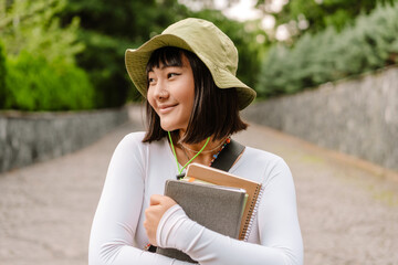 Young asian woman smiling and holding exercise books while walking at park