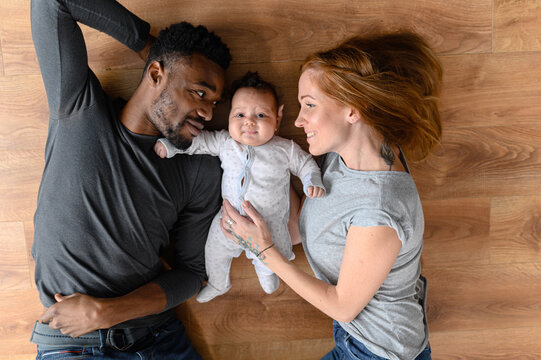 Happy mixed-race international family of three lying on the floor, happy black husband, caucasian wife and their biracial baby girl looking at the camera, top view