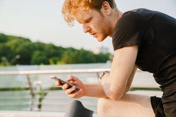 Ginger sportsman using mobile phone while working out on bridge