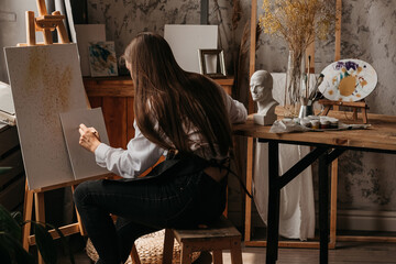 A young handsome artist writes with pencil on canvas in a creative workshop and sits next to art supplies lying on a small wooden table.