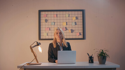 Businesswoman looking at camera near laptop and lamp on office table