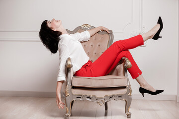 Studio portrait of attractive young woman with dark hair in white shirt and red pants sitting on...