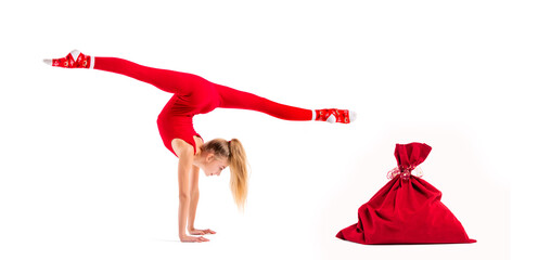 Slender girl gymnast in red tight-fitting suit and santa claus hat posing next to bag for gifts...