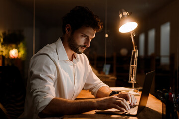 Brunette man wearing shirt working with laptop while sitting at desk