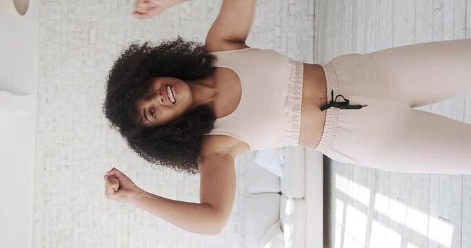 Trendy happy black mixed race woman vlogger listeng music, dances and recording videostream video for social media at home. Creating dancing video using tiktok app. Vertical video.