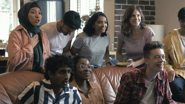 Young multicultural friends students sitting on sofa and watching sport match on TV. They throwing arms up in excitement and yelling while celebrating goal