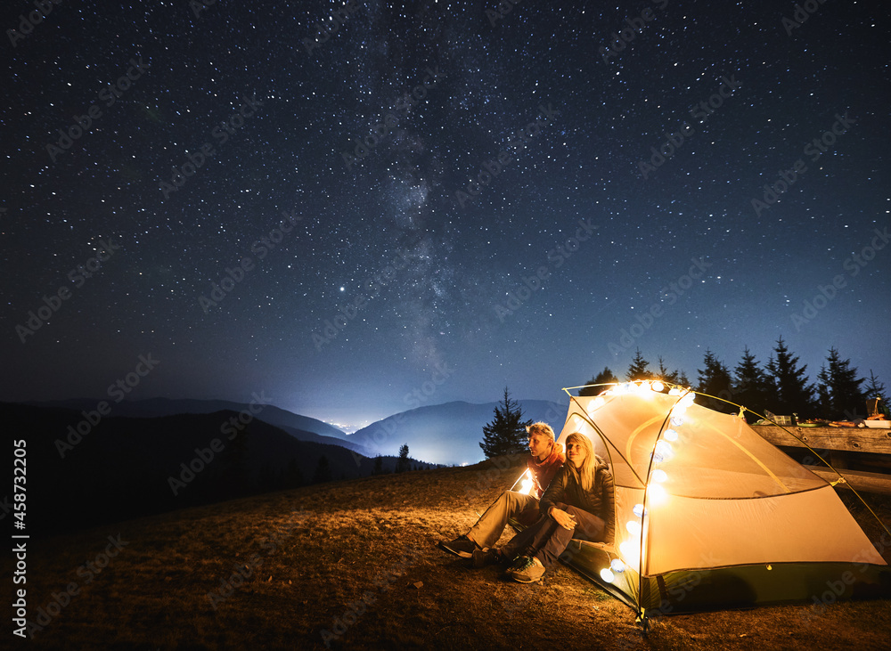 Wall mural couple sitting down in illuminated tent relaxing after long day of hiking and enjoying the view of e