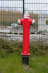 Fototapeta na wymiar Roter Hydrant zum Feuer löschen steht auf einer Wiese vor einem grünen Zaun.