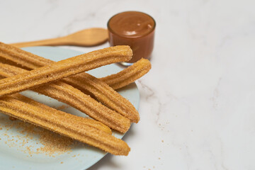 Traditional churros with sugar and cinnamon served on a marble table, with a bowl of caramel.