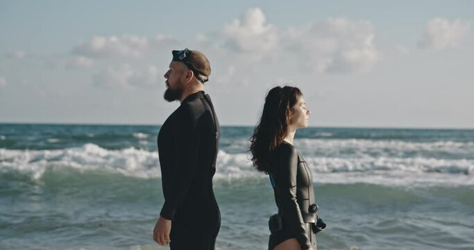 Couple of man and woman in swimming suits. Spectacular couple standing against sea waves in black suits. Brunette with long hair covering her eyes, man looking into the distance. Concept of cinema