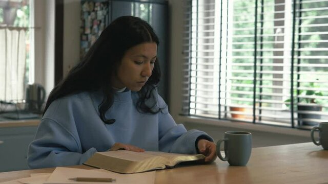 Young female student study at home in the morning drinking tea or coffee. Christian hispanic girl with black hair reading holy book, bible. Devotional