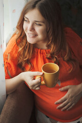 beautiful pregnant young woman sitting in kitchen near window with cup of hot tea drink, motherhood concept