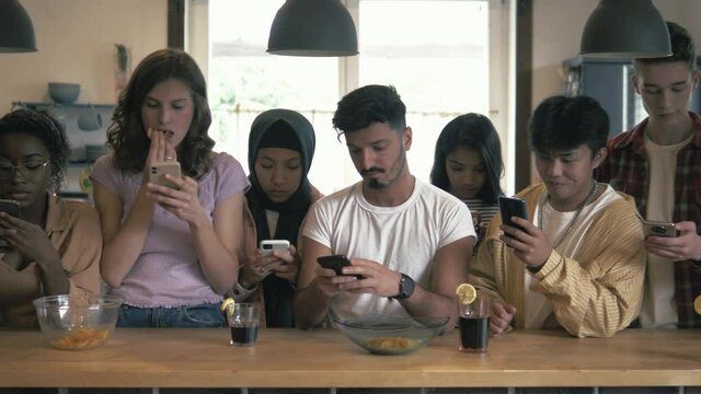 Group of young addicted multicultural friends phubbing  at home. Busy bored teenagers using smart phones with disinterest on each other. Technology and smart phone addiction.