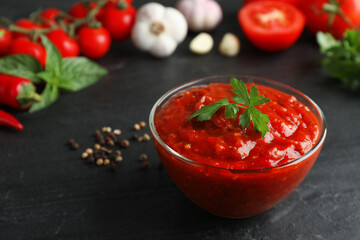 Delicious adjika sauce with parsley in glass bowl and ingredients on black table, closeup