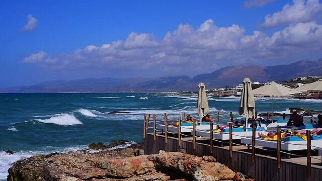 HERSONISOS, GREECE - Aug 11, 2021: A windy day on the beach in Hersonissos, Greece, and tourists in sunbeds next to a rock formation.