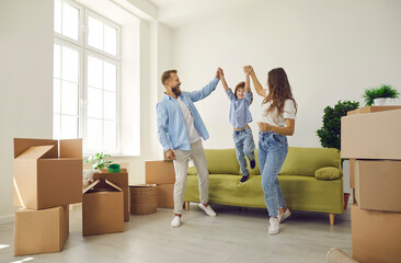 Fototapeta na wymiar Happy family playing and having fun in new home full of cardboard boxes. Cheerful mom and dad holding son by the hands as he jumps off sofa in living room. Real estate, buying and moving house concept