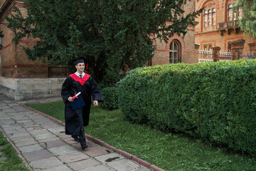 Fototapeta na wymiar Happy male graduate walking in university campus in graduation robe, copy space for text