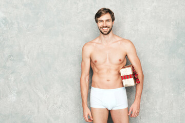 Obraz premium Sporty handsome strong man. Healthy athletic fitness model posing near grey wall in white underwear. Confident sexy fashion male with naked nude torso with present. Lambersexual holding gift box