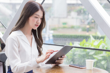 Young Asian business woman is looking at tablet or notepad in her hands while she is sitting at coffee shop among business background in the city.