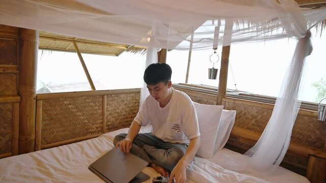Asian man working with laptop and taking a photo with retro camera in wooden thatched resort among the nature