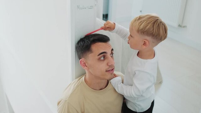 A little child measuring height of his father using pencil standing near wall at home