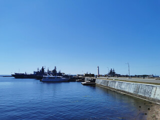 Fototapeta na wymiar View of warships, boats and a pier with a lighthouse from Petrovskaya Embankment against the blue sky on a summer day in Kronstadt.