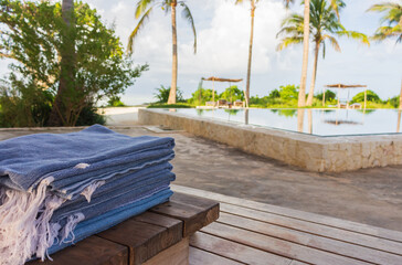Stack of clean towels on wooden deck near swimming pool with palm trees. Lounge zone in tropical resort hotel. Empty hotel patio. Leisure and relax background. Beach towel in luxury resort. 