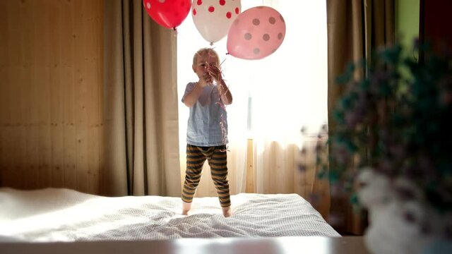 Cute little boy in striped clothes jumping on bed in cozy sunny bedroom. Child play with balloons at home enjoy carefree time. Kid have fun and laughing on quarantine, coronavirus home isolation.