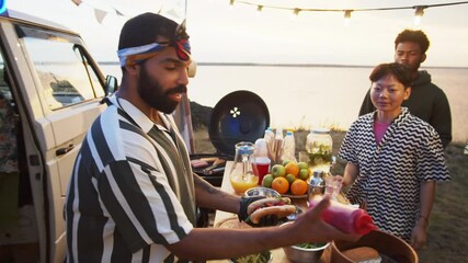 Cheerful food truck seller adding ketchup to hot dog, giving it to young Asian woman and showing thumbs up while working on summer festival