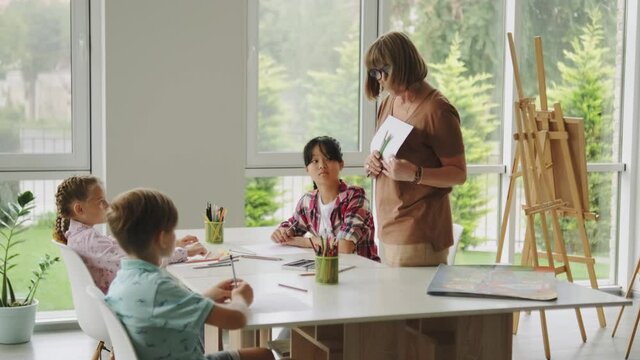 A woman teacher explain for children task of drawing holding paper and pencils in the art school