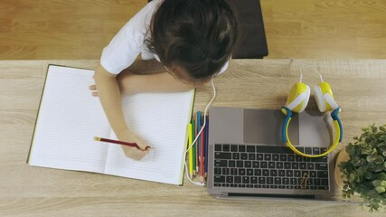 Top view 4K of adorable asian girl kid, wearing headphone and writing on paper book, is learning online from computer laptop on wooden desk shows concept of home education using internet technology.