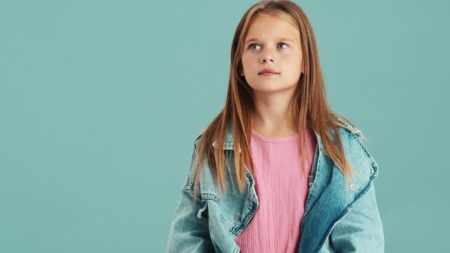 A Confused Small Girl Wearing Jeans Jacket Shrugging Shoulders Standing Isolated Over A Blue Wall In The Studio