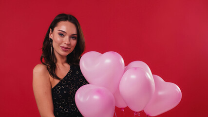 cheerful woman in black dress holding pink balloons isolated on red.