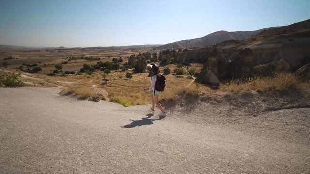 Traveler girl walking, back view with backpack and leather hat, tourist. Fairy chimneys park in the background. rock formations. Travel and vacation, tour Goreme, Cappadocia, Turkey.