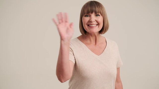 A smiling cheerful mature woman with bob hairstyle showing hello gesture at the camera on gray-beige background in studio