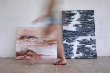 Contemporary art. Selective focus on two abstract and colorful paintings, laying against the wall. A blurred young caucasian woman walks across the room.