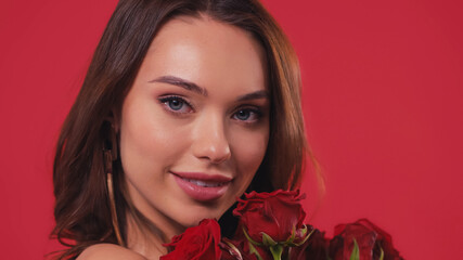 pleased young woman smiling near roses isolated on red.